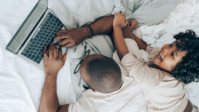A father works on a laptop in bed while his child laughs beside him