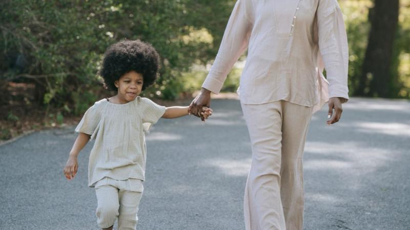 A mother and her young son walk together hand in hand along a sunlit path