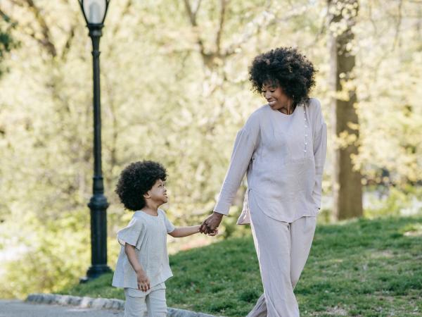 A mother and her young son walk hand in hand along a tree-lined path, sharing a smile.