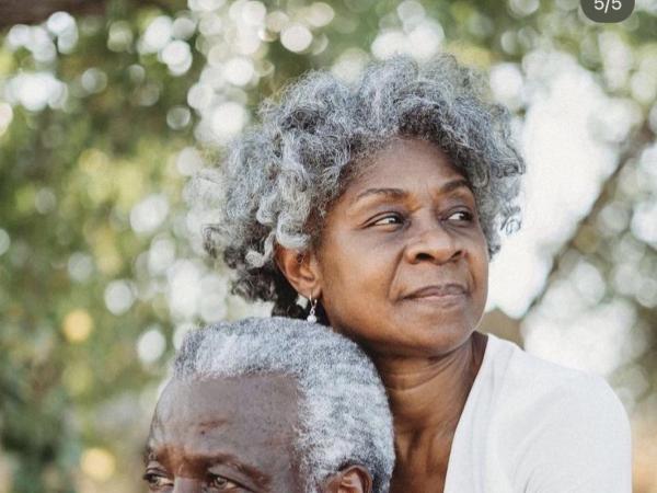 An elderly couple embrace outdoors, looking to the horizon